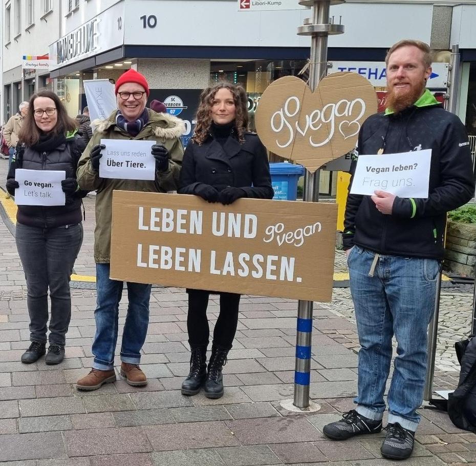 Vier Menschen (zwei Männer, zwei Frauen) stehen warm angezogen in einer Fußgängerzone. Ganz links eine Frau mit dunkler Kleidung und mittellangen Haare. Daneben ein Mann mit rotem Beanie und grünem Parka, daneben eine Frau mit dunkler Kleidung und Locken und daneben ein Mann mit Bart und blauer Kleidung. Sie halten Schilder mit der Aufschrift "Leben und vegan leben lassen", "Go Vegan - let's talk", "Lass uns reden - über Tiere" und "Vegan leben - frag uns".